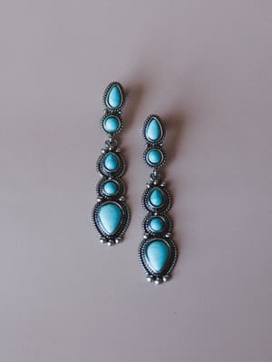 A pair of multi-stone turquoise drop earrings with oxidized silver rope-detail bezels, laid flat against a soft taupe background — the kind of earrings that do all the talking.