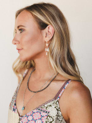 A close profile shot of a model wearing layered seashell and star charm drop earrings in gold, styled with a mixed floral print tank and layered necklaces — the whole look has that easy, sun-warmed boho energy.