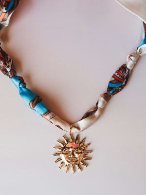 A close-up of a braided fabric cord necklace in a multicolor printed satin with a bold gold sun face pendant — the kind of piece that looks like it has a story already.
