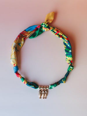 A colorful printed fabric charm necklace laid flat against a soft blush background — the tropical multiprint scarf-style band is tied at the top and anchored by a small gold fish charm pendant, giving it that collected, well-traveled energy.