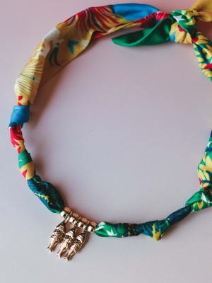 A colorful tropical-print fabric necklace laid flat on a soft pink surface, finished with a gold figural charm — the kind of piece that feels like a vacation souvenir you'd actually wear again.