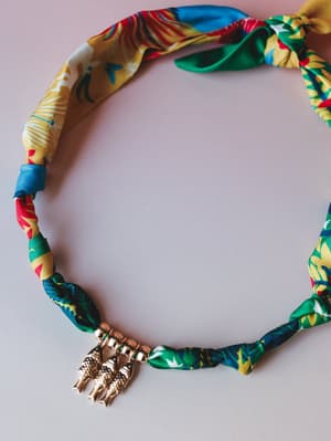 A colorful tropical-print fabric necklace laid flat on a soft pink surface, finished with a gold figural charm — the kind of piece that feels like a vacation souvenir you'd actually wear again.