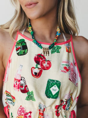 A close-up torso shot of a whimsical novelty-print halter top styled with a knotted scarf-and-chain charm necklace featuring gold fish pendants — playful, warm, and full of personality.