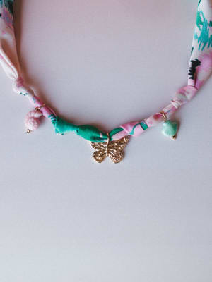 A whimsical fabric-cord necklace with a gold butterfly pendant, tiny ceramic charms, and a twisted floral-print ribbon in pink and green — laid flat against a soft lavender-grey surface.