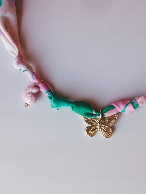 A close-up of a fabric-cord necklace in twisted pink floral and green fabric, anchored by a gold butterfly charm and a small pink shell bead — playful, handcrafted-feeling, and full of personality.