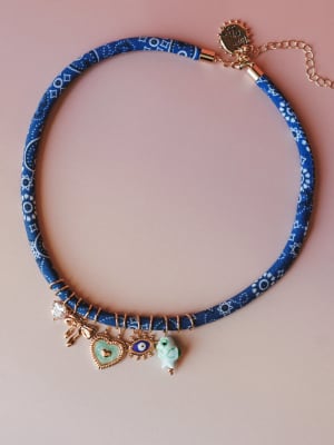 A fabric-wrapped blue bandana-print choker necklace laid flat against a blush background, adorned with an eclectic mix of gold charms — a crystal bow, enamel heart, evil eye, and ceramic fish bead — that feels like a little collection of lucky things