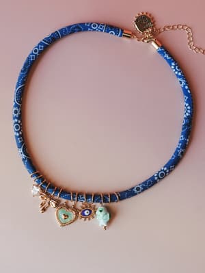A fabric-wrapped blue bandana-print choker necklace laid flat against a blush background, adorned with an eclectic mix of gold charms — a crystal bow, enamel heart, evil eye, and ceramic fish bead — that feels like a little collection of lucky things