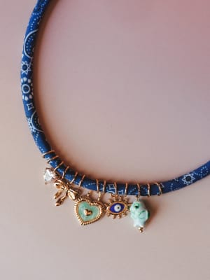 A fabric-wrapped navy choker necklace with a playful cluster of gold charms — a crystal drop, bow, enamel heart, evil eye, and floral ceramic bead — laid flat on a soft blush background.