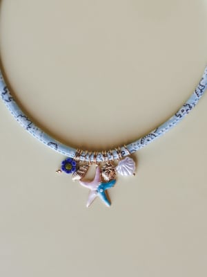 A fabric-wrapped choker necklace in light blue with a tiny printed pattern, hung with a cluster of ocean-themed charms — a starfish, seashell, conch, flower bead, and rose gold accents — shot flat against a warm neutral surface.