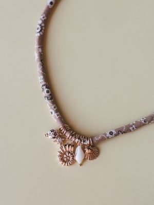A fabric-wrapped choker necklace in dusty mauve with a ditsy floral print, adorned with a cluster of rose gold sea-inspired charms — a scallop shell, sunflower shell, teardrop bead, starfish, and painted ceramic bead — laid flat on a warm neutral sur