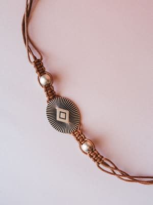 A close-up of a brown macramé cord belt with a engraved sunburst medallion centerpiece and silver beaded accents, shot flat against a soft blush background — the kind of detail that makes a simple dress feel intentional.