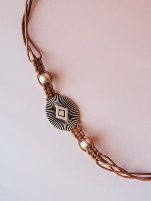 A close-up of a brown macramé cord belt with a engraved sunburst medallion centerpiece and silver beaded accents, shot flat against a soft blush background — the kind of detail that makes a simple dress feel intentional.