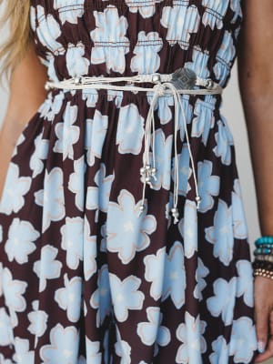 A close-up torso shot of a floral smocked dress styled with a cream macramé belt featuring a silver concho centerpiece and beaded tassel ties — the kind of detail that turns a great dress into a whole moment.