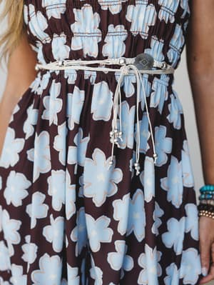 A close-up torso shot of a floral smocked dress styled with a cream macramé belt featuring a silver concho centerpiece and beaded tassel ties — the kind of detail that turns a great dress into a whole moment.