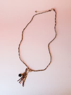 A delicate twisted chain necklace laid flat on a blush pink surface, its pendant a playful cluster of earth-toned seed beads trailing downward like fringe — earthy, a little whimsical, the kind of piece you forget you're wearing until someone asks ab
