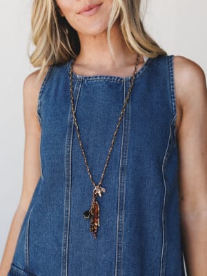A long twisted rope chain necklace with a dangling charm cluster of brown beads, amber stones, and mixed pendants, styled against a sleeveless denim top for a casual boho feel.