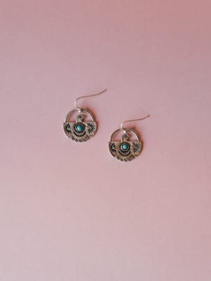 A pair of oxidized silver thunderbird drop earrings with turquoise stone centers, laid flat against a soft dusty rose background — earthy, storied, and the kind of thing you reach for every single day.