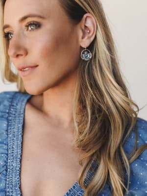 A close-up portrait shot showing silver thunderbird drop earrings with a turquoise stone center, worn against soft natural light — earthy, expressive, and quietly striking against a denim blue smocked top.