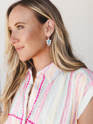 A close-up portrait of a model wearing silver thunderbird drop earrings with a turquoise stone center, paired with a multicolor stripe button-down top — the whole look has an easy, sun-warmed southwestern feel.