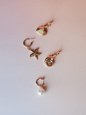Four individual gold earrings laid out on a soft blush-to-lavender gradient surface — a clam shell, a starfish, a spiral shell, and a pearl drop hoop, each one feeling like a little keepsake from a really good beach trip.