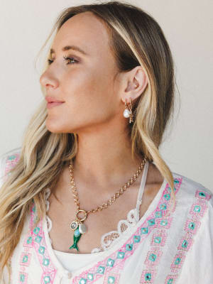 A close portrait of a model wearing layered gold jewelry — pearl and starfish charm hoop earrings paired with a chunky gold chain necklace featuring mermaid tail, peace sign, and moonstone charms — styled with a white embroidered top for an effortles