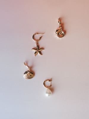 Four mismatched gold charm earrings laid out separately on a soft blush-to-lavender gradient surface — a starfish hoop, spiral shell, clam shell, and pearl drop hoop, each with their own personality but clearly made to live together.