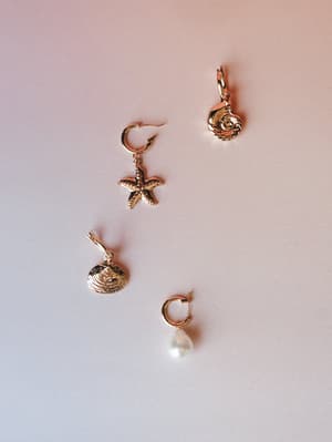 Four mismatched gold charm earrings laid out separately on a soft blush-to-lavender gradient surface — a starfish hoop, spiral shell, clam shell, and pearl drop hoop, each with their own personality but clearly made to live together.