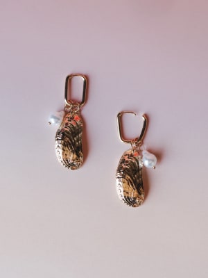 A pair of statement drop earrings featuring gold-dipped shell charms with natural swirling texture, small freshwater pearl drops, and rectangular gold huggie hoops — laid flat on a soft blush background.