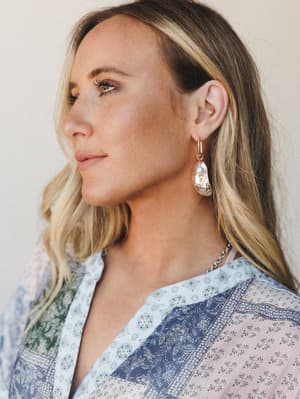 A close profile shot highlighting gold drop earrings with a hammered medallion and pearl detail, styled against a patchwork-print boho top in soft blues and blush — the whole thing feels like a Sunday that got unexpectedly beautiful.
