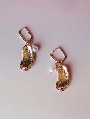 A pair of gold shell-shaped drop earrings with pearl accents, laid flat against a soft blush background — the kind of earrings that feel equally at home at a beach wedding or a Thursday dinner out.