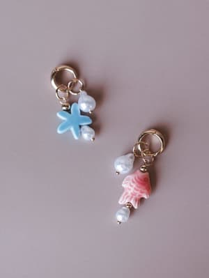 A mismatched pair of gold hoop charm earrings laid flat on a soft mauve background — one dangling a sky blue starfish charm with pearl beads, the other a pink fish charm with pearl beads, both playful and ocean-inspired.