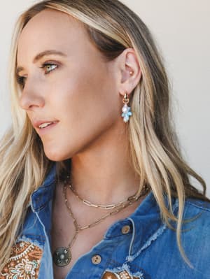 A close-up portrait of a blonde model in profile, styled with gold hoop earrings featuring blue flower and pearl charms, layered gold chain necklaces, and a distressed denim jacket over a printed top — warm, effortless, and quietly put-together.