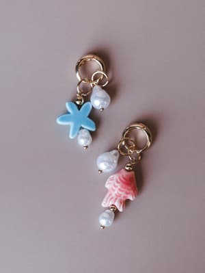 A mismatched pair of ocean-inspired charm earrings — one with a powder blue starfish, one with a painted pink shell — both dangling from gold huggie hoops with freshwater pearl accents, shot flat against a soft mauve background.