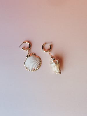A mismatched pair of shell drop earrings — one scallop, one conch — hanging from small gold hoops and laid flat on a dusty rose gradient background, with a dreamy, seaside-collected feel.