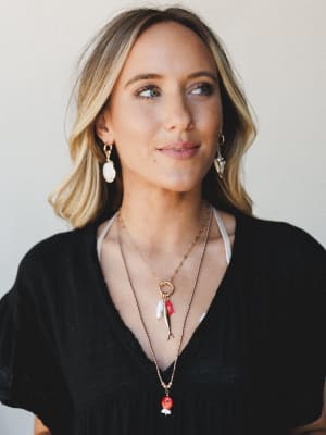 A close portrait of a model wearing layered gold charm necklaces and mismatched shell drop earrings against a soft neutral backdrop — the kind of jewelry stack that looks collected over time, not bought all at once.