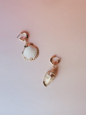 A mismatched pair of seashell drop earrings — one scallop, one spiral — both gold-rimmed and hung from small gold hoops, shot flat against a soft blush backdrop with a dreamy, coastal ease.