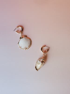 A mismatched pair of seashell drop earrings — one scallop, one spiral — both gold-rimmed and hung from small gold hoops, shot flat against a soft blush backdrop with a dreamy, coastal ease.