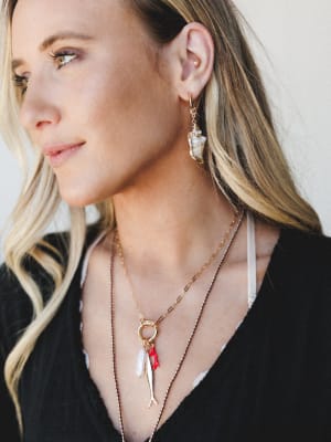 A close-up lifestyle shot of a model wearing layered gold jewelry — a seashell drop earring and a charm necklace stack with pearl, fish, and red enamel pendants — against a soft neutral background, with a calm, coastal-romantic feel.