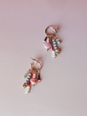A pair of gold hoop earrings layered with mismatched beaded charms — shell-shaped beads, seed beads, and tube beads in blush, sky blue, magenta, navy, and cream — laid loosely against a soft dusty pink background.
