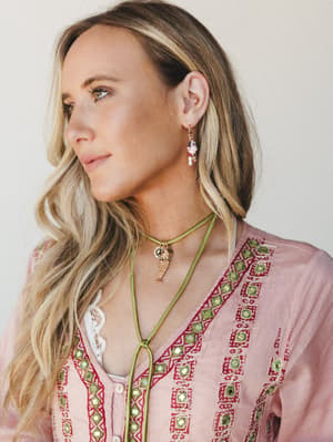 A close-up lifestyle shot of a model in a blush embroidered top wearing layered boho jewelry — beaded dangle earrings and a green leather cord charm necklace — with a soft, dreamy studio feel.