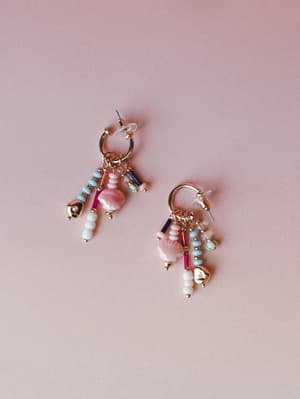 A pair of gold hoop earrings with playful multi-charm dangles — shell beads, seed beads, and glass tubes in pink, sky blue, and fuchsia — shot flat against a soft dusty rose background that makes the whole thing feel like a handful of summer memories