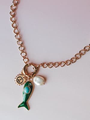 A gold rolo chain necklace styled with three charms — an enamel green fish with a turquoise stone eye, a freshwater baroque pearl drop, and a small gold sun peace sign medallion — shot flat against a soft lavender-grey background.
