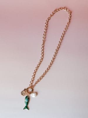 A gold rolo chain charm necklace laid flat against a soft pink-to-white gradient background, anchored by a playful cluster of charms — an enamel green fish with a turquoise stone, a freshwater pearl drop, and a small peace sign medallion.