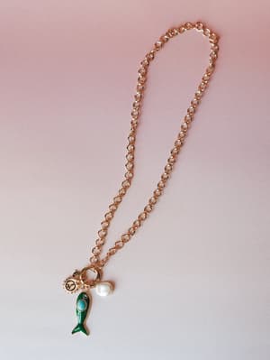 A gold rolo chain charm necklace laid flat against a soft pink-to-white gradient background, anchored by a playful cluster of charms — an enamel green fish with a turquoise stone, a freshwater pearl drop, and a small peace sign medallion.