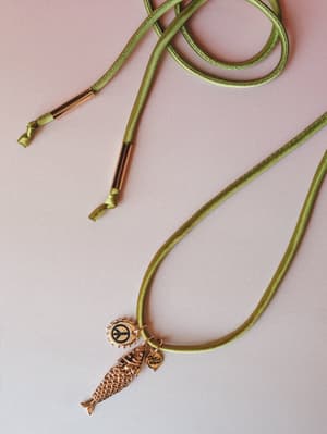 A moss green satin wrap necklace laid flat against a soft pink background, its rose gold charm cluster — a peace sign, articulated koi fish, and branded coin — doing all the personality work.