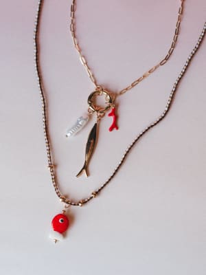 Two layered necklaces styled together on a soft blush background — a braided cord style with a red glass fish pendant and gold beads, alongside a gold paperclip chain with a charm ring holding a gold fish, red coral branch, and pearl drop charms.