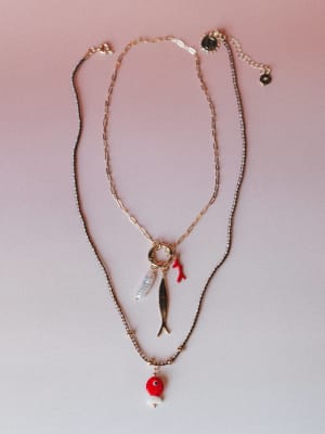 A layered necklace set styled flat against a soft pink backdrop — one gold paperclip chain with a charm cluster (fish, coral branch, and pearl drop) and one delicate dark beaded chain ending in a red enamel fish bead and pearl drop, designed to wear
