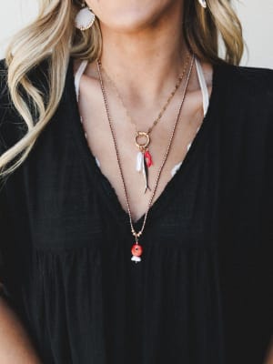 A close-up lifestyle shot of layered gold necklaces styled against a black v-neck top — the stack features a charm necklace with a red coral branch, baroque pearl, and fish pendant, plus a long beaded chain with a red fish bead drop, all paired with