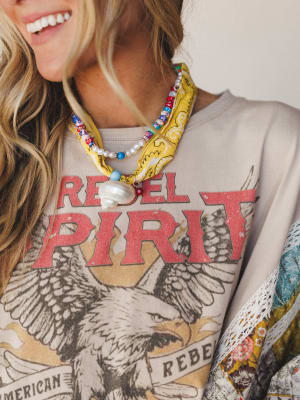 A close-up shot of layered boho accessories — a colorful beaded and pearl necklace with a shell charm sits over a yellow bandana scarf tied loosely at the neck, all styled over a washed graphic sweatshirt with a rebel eagle motif and a peek of floral