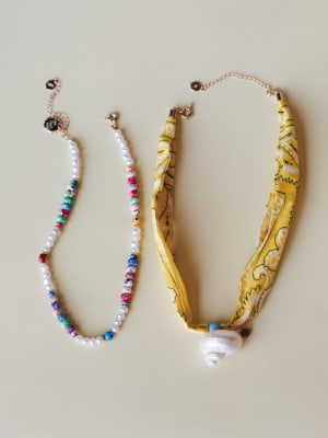 Two boho-spirited necklaces laid flat on a warm neutral surface — a colorful mixed bead and freshwater pearl strand alongside a yellow bandana-print fabric necklace finished with a swirled shell pendant and gold chain closure.
